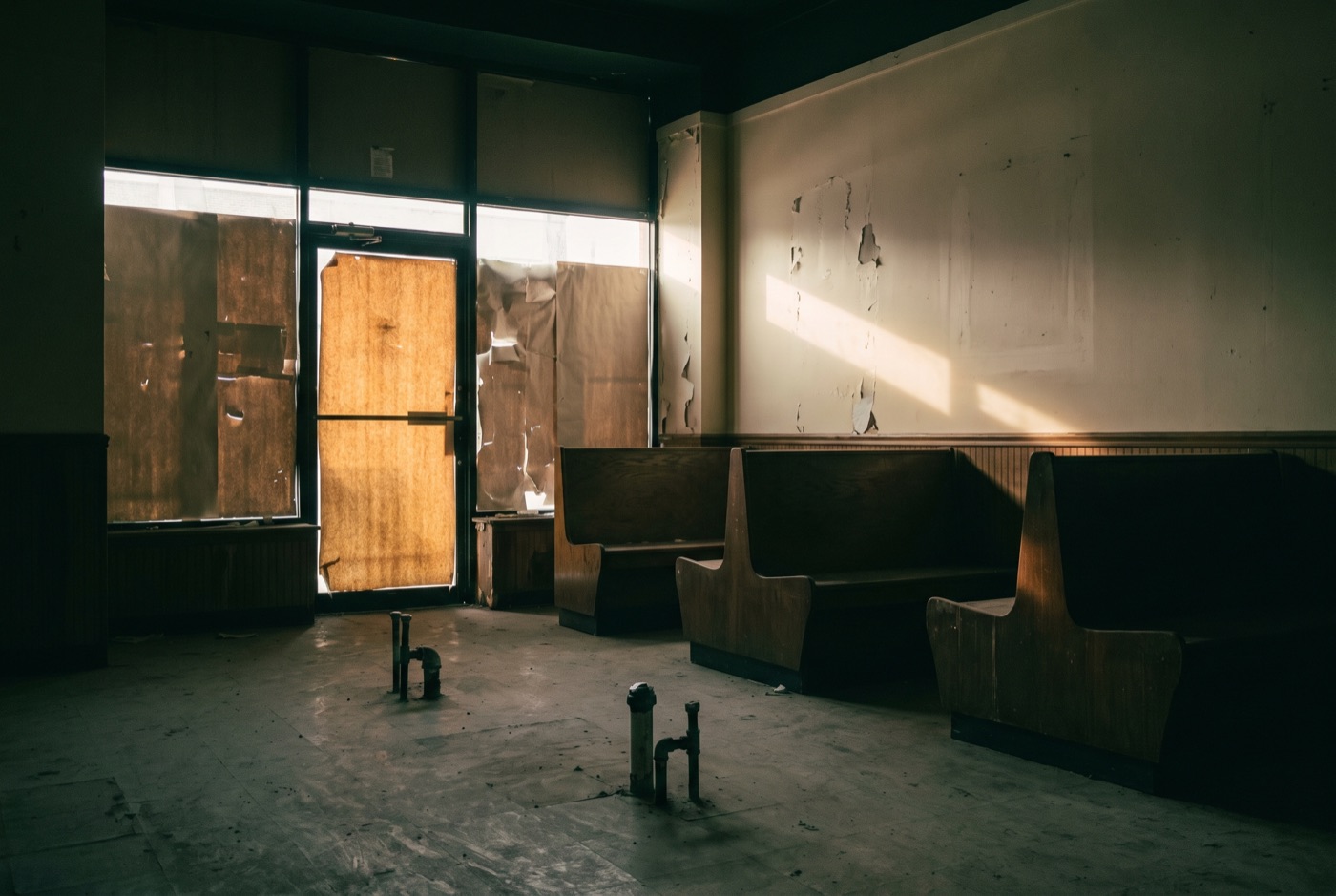 A dark, stripped second-generation restaurant interior with capped plumbing stubs and a single shaft of daylight