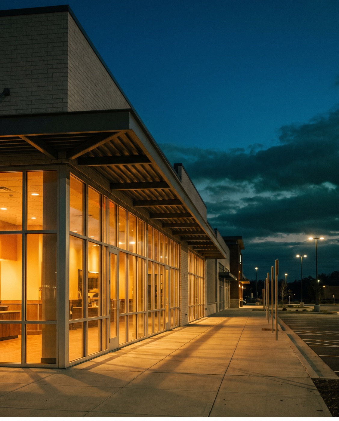 Dusk exterior of a modern retail-center endcap restaurant with warm light spilling onto the sidewalk