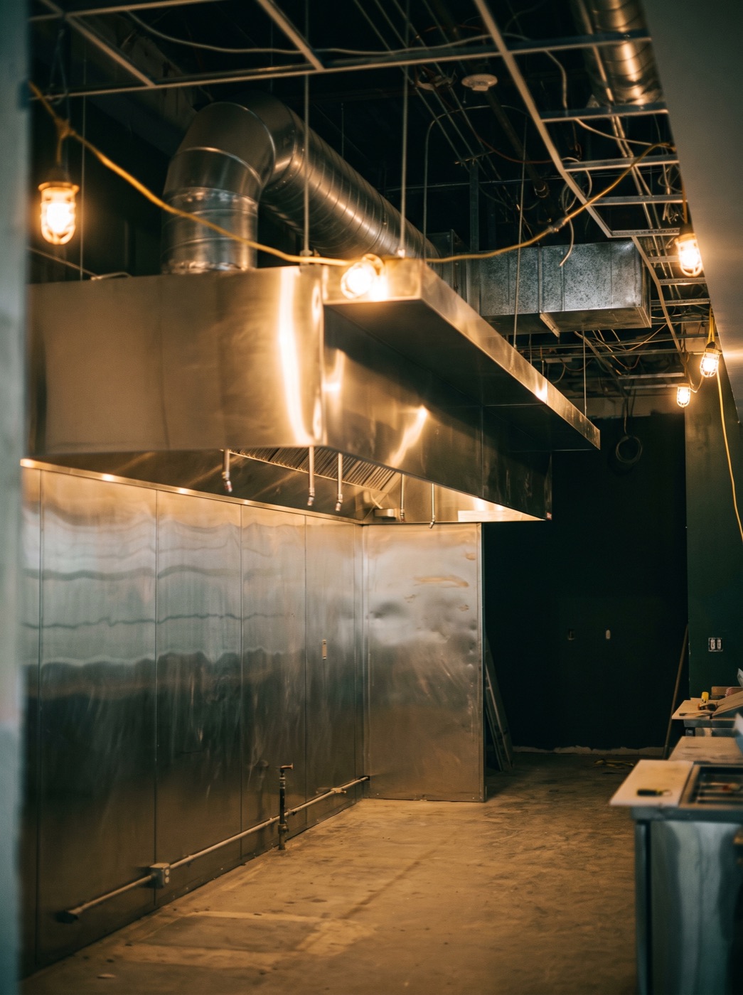 A stainless Type I exhaust hood above an empty cook line with ductwork rising into an open ceiling
